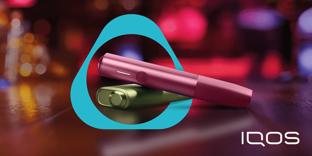 Immagine con dispositivi IQOS ILUMA ONE uno sovrapposto sull'altro di colore verde e fucsia poggiati su un piano di legno. Sfondo con delle luci soffuse viola e logo iqos di colore turchese al centro che avvolge i dispositivi