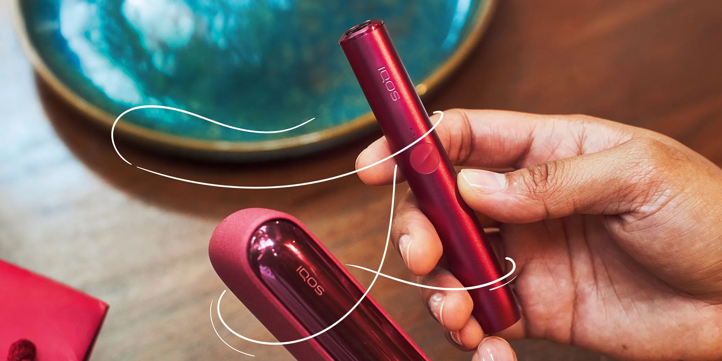 Mano che tiene un dispositivo IQOS ILUMA di colore Sunset Red su un tavolo di legno con un copy di colore bianco Un passo in più