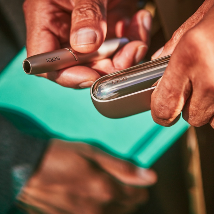 Un IQOS brilliant gold nella mano destra di un uomo, il charger nella sinistra