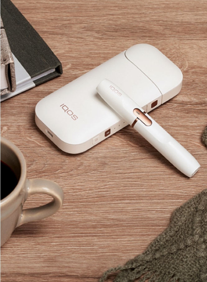 White IQOS 2.4 PLUS holder and charger on a wooden table next to a cup