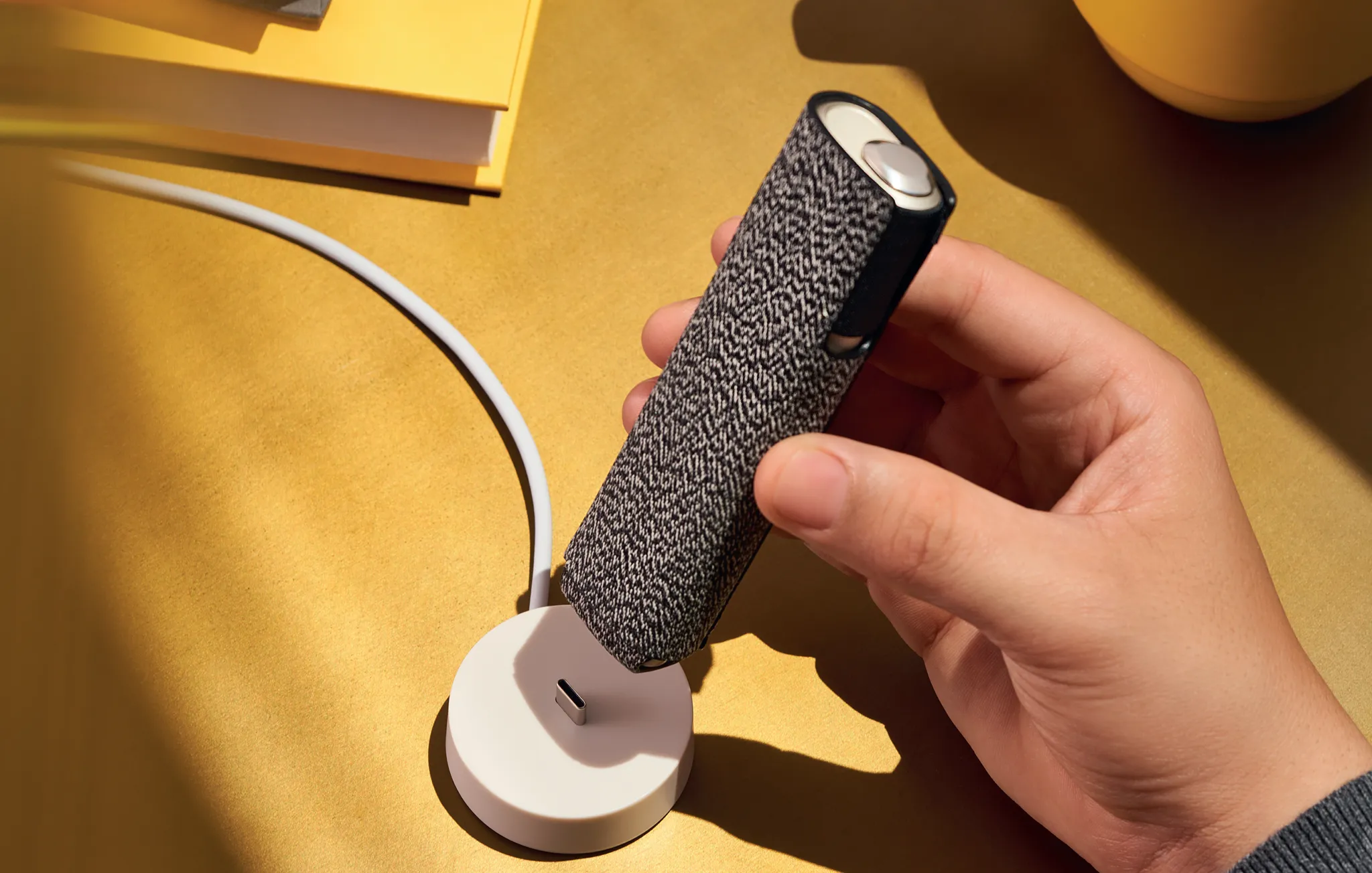 Mano che mette in carica un dispositivo IQOS ILUMA ONE con Cover Plus nera e bianca, su un tavolo giallo.