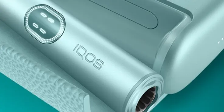 Garnet Red IQOS ILUMA i PRIME device and its charger on a brown notebook