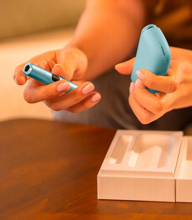 Hands holding turquoise IQOS device and charger over pink packaging on wooden table