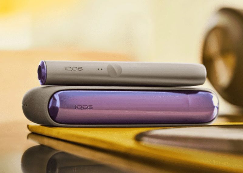 A hand holding an IQOS ILUMA device and a charger on a table.