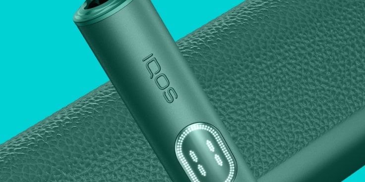 IQOS ILUMA i PRIME nel colore verde con holder con schermo touch e caricatore su sfondo turchese